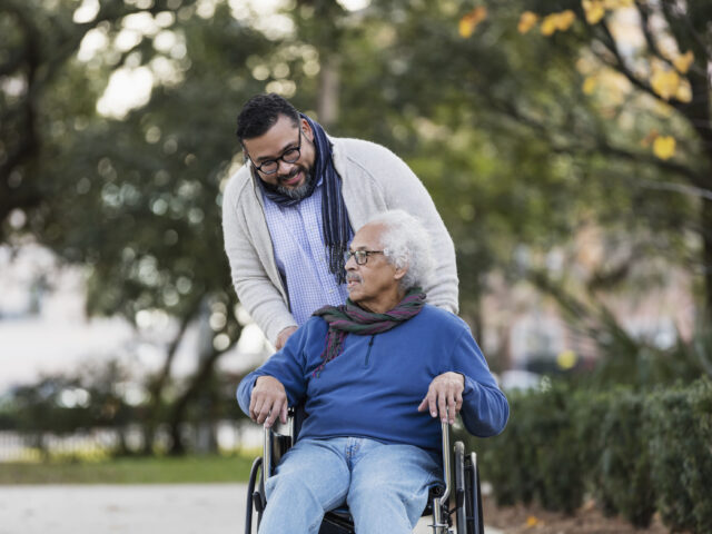 A caregiver accompanying an older man in a wheelchair outdoors.