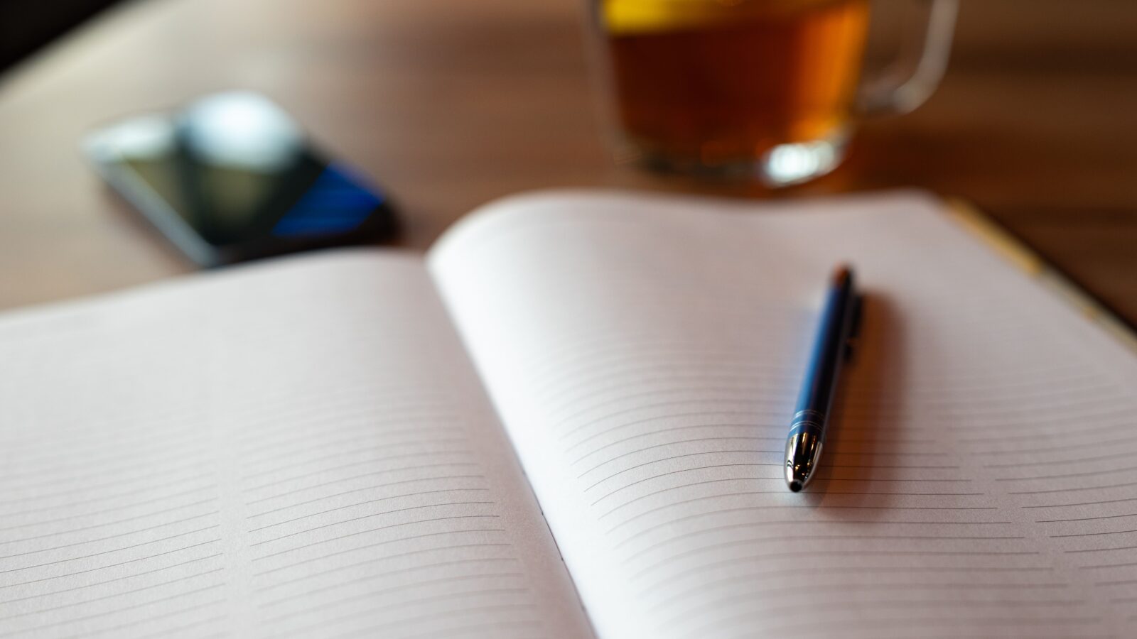 An open notebook and pen beside a cup of tea on a wooden table.