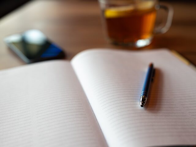 An open notebook and pen beside a cup of tea on a wooden table.